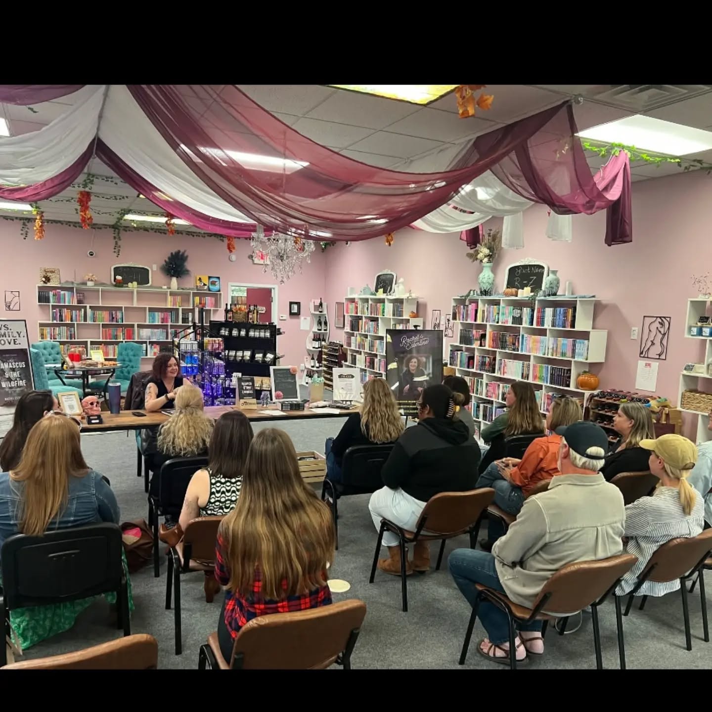 People seated in a room with bookshelves, listening to a speaker at a table, decorated with drapes and hanging ornaments.