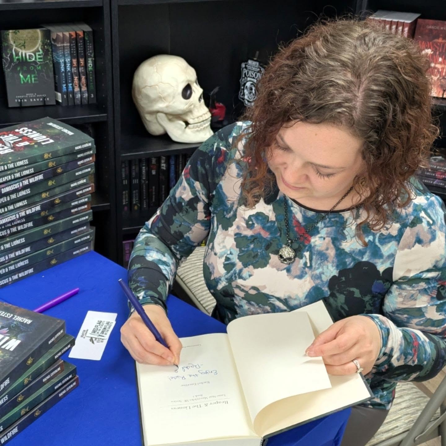 Woman writing in a book at a table with bookshelves and a skull decoration behind her.