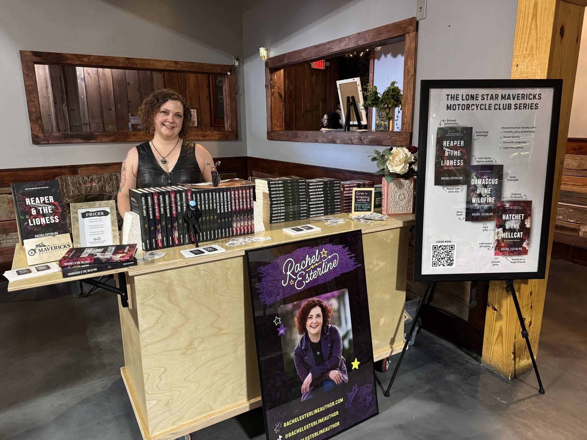 Woman standing behind a table with books and promotional materials, in a room with wooden accents and a framed poster on an easel.