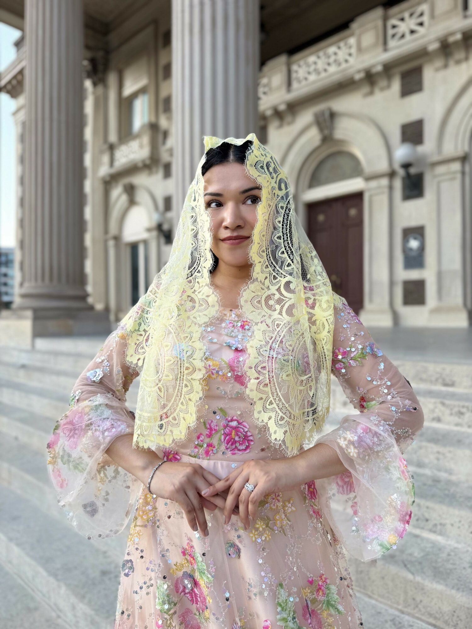 Woman wearing a lace headscarf and traditional dress standing on steps in front of a historic building with columns.