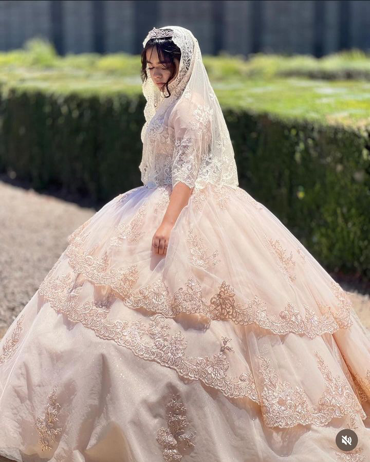 Person in a wedding dress with lace veil outdoors, hedge and trees in background.