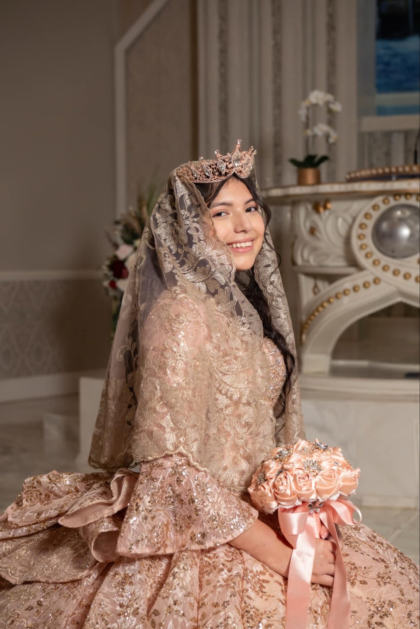 Woman in ornate dress with lace veil and tiara, smiling, sitting indoors with elegant decor in background.