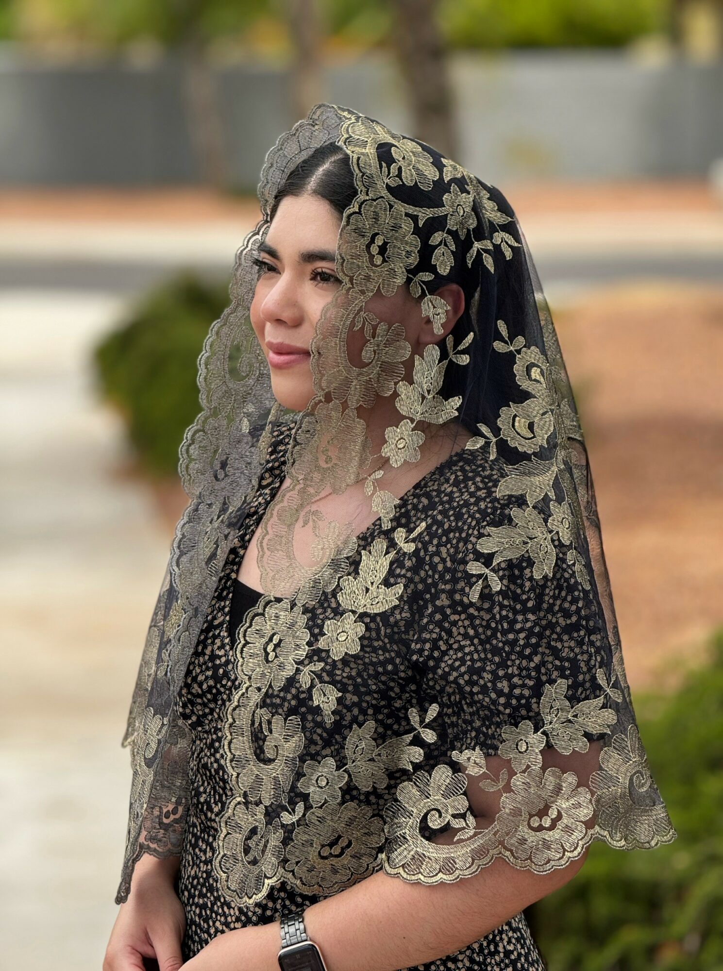 Woman wearing a black and gold embroidered veil outdoors, with trees and a pathway in the background.