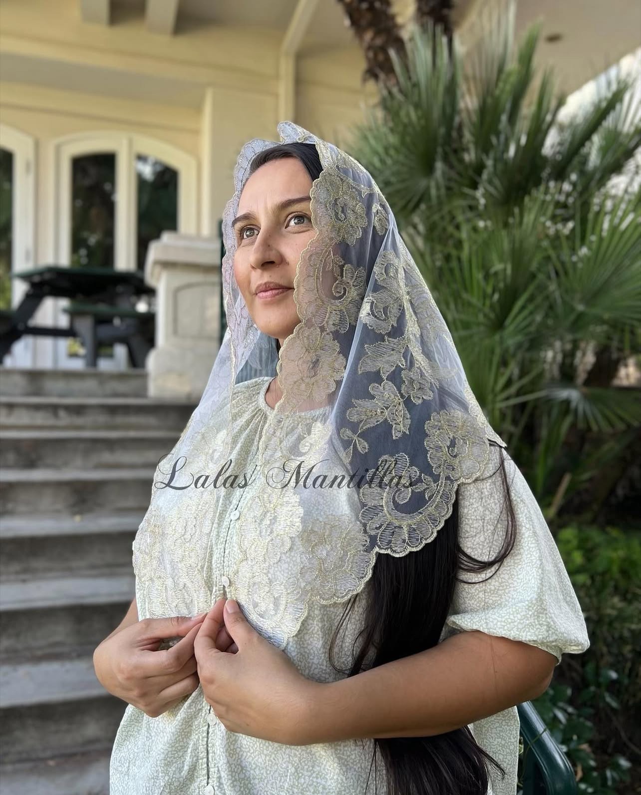 Woman wearing lace veil and white dress, standing outdoors near stairs and greenery.