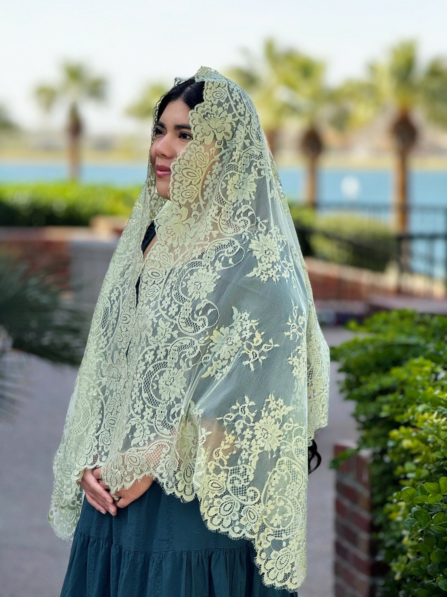 Woman wearing a lace veil outdoors with trees and a body of water in the background.