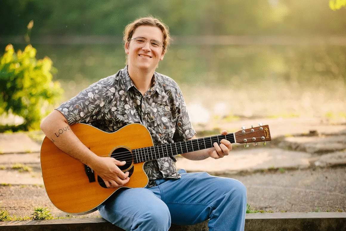 Person sitting outdoors playing an acoustic guitar, smiling, with a lake and trees in the background.
