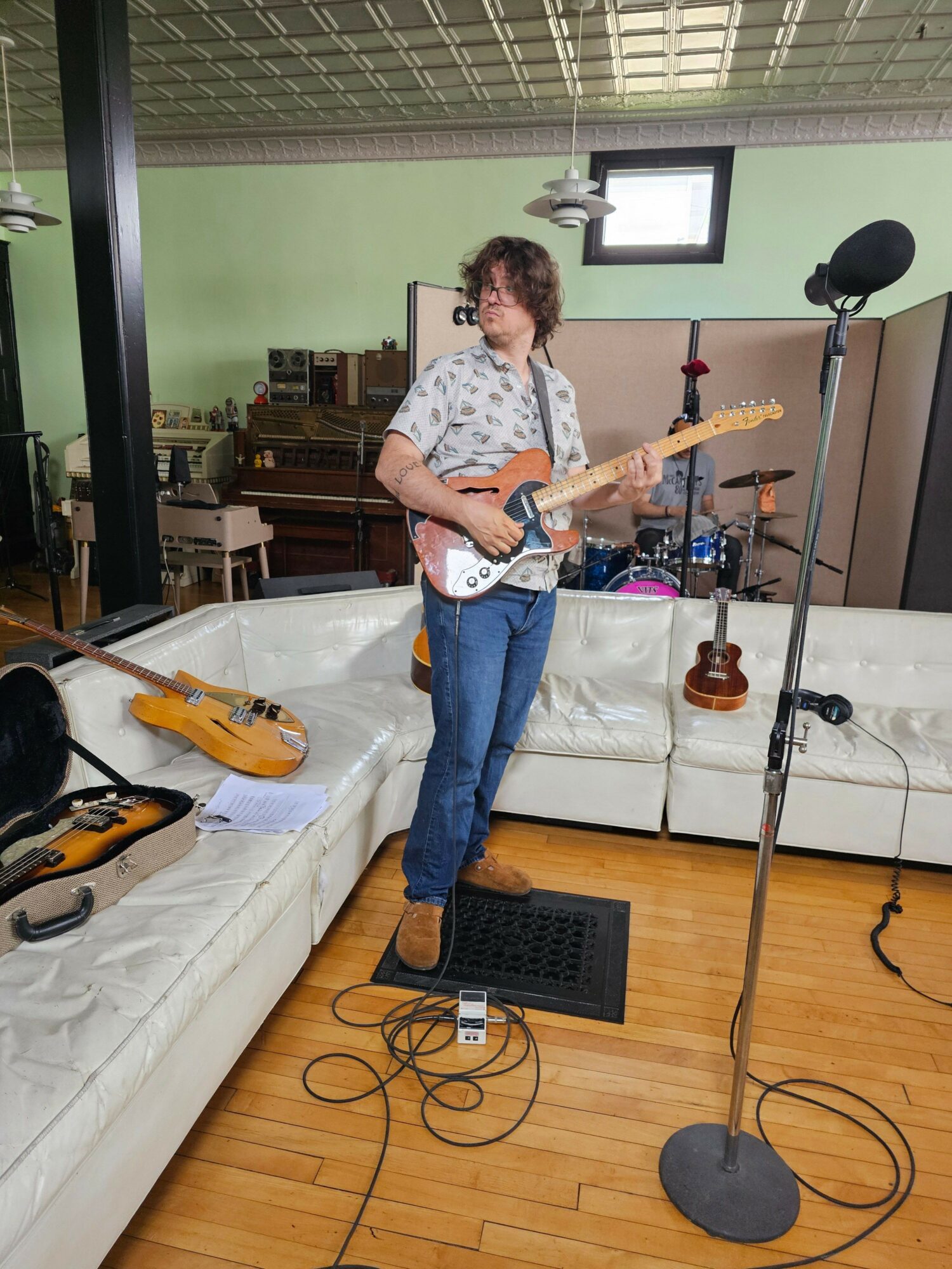 Person playing an electric guitar in a room with recording equipment and microphones, standing on a wooden floor.