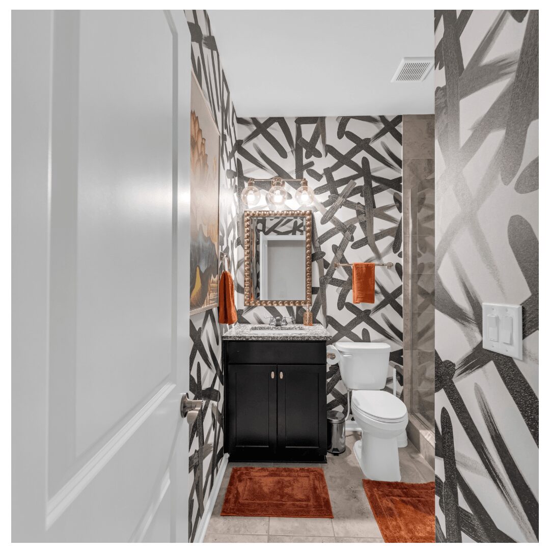 Bathroom with black and white patterned walls, mirror, sink, toilet, and orange accents, including a rug and towel.