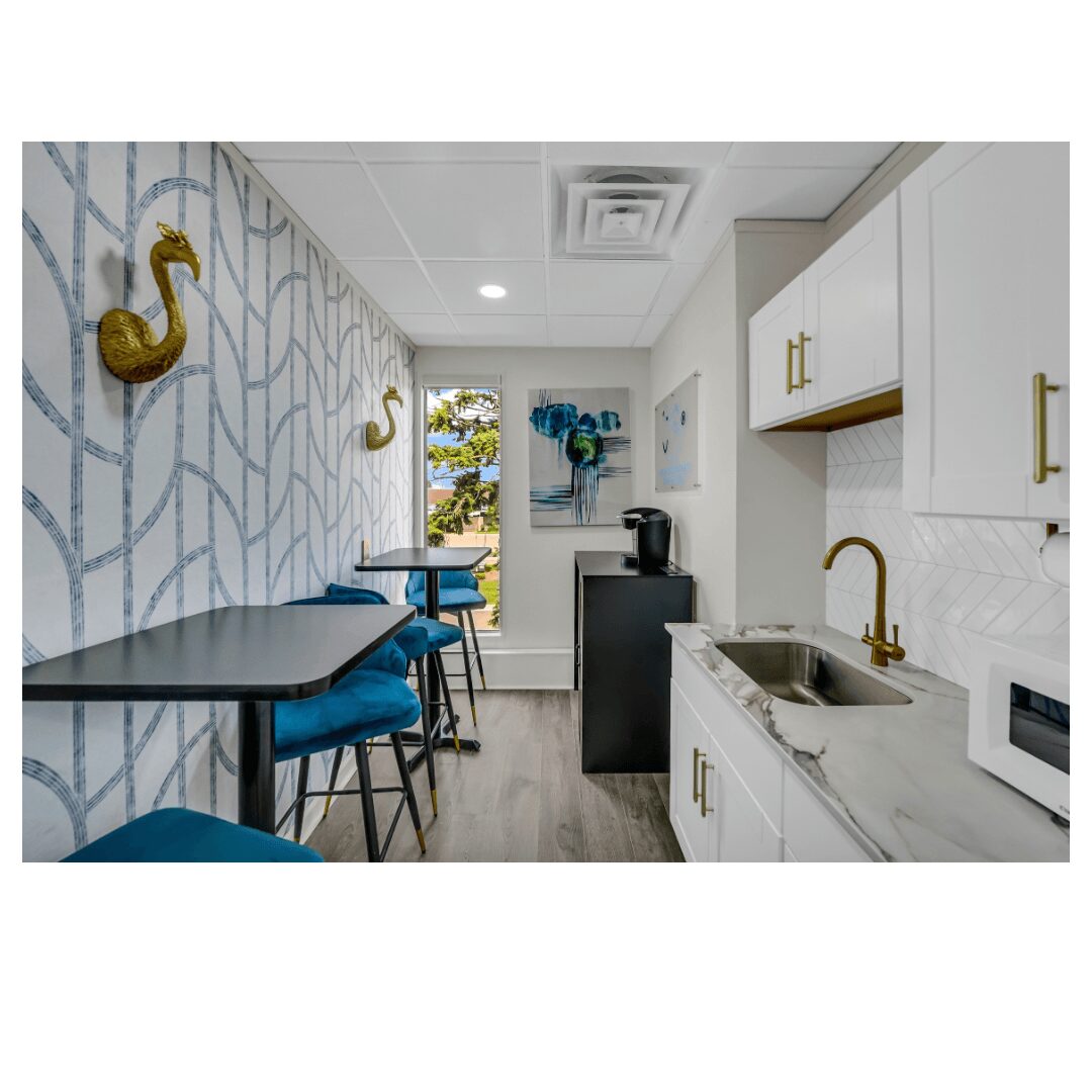 Kitchen with white cabinets, gold hardware, a sink, and a small dining area with blue chairs, window view outside.