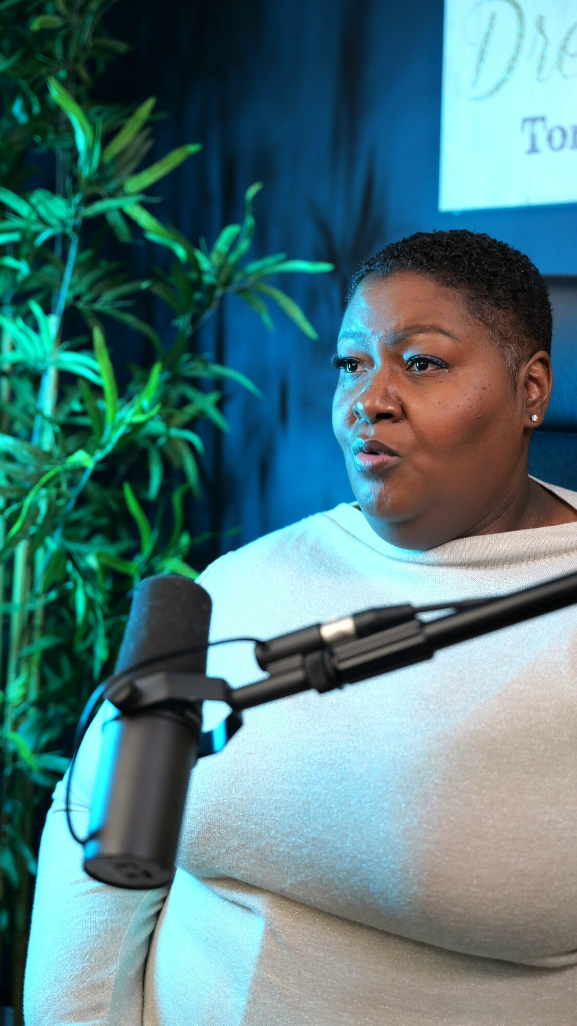 Woman speaking into a microphone with a plant and a screen in the background.