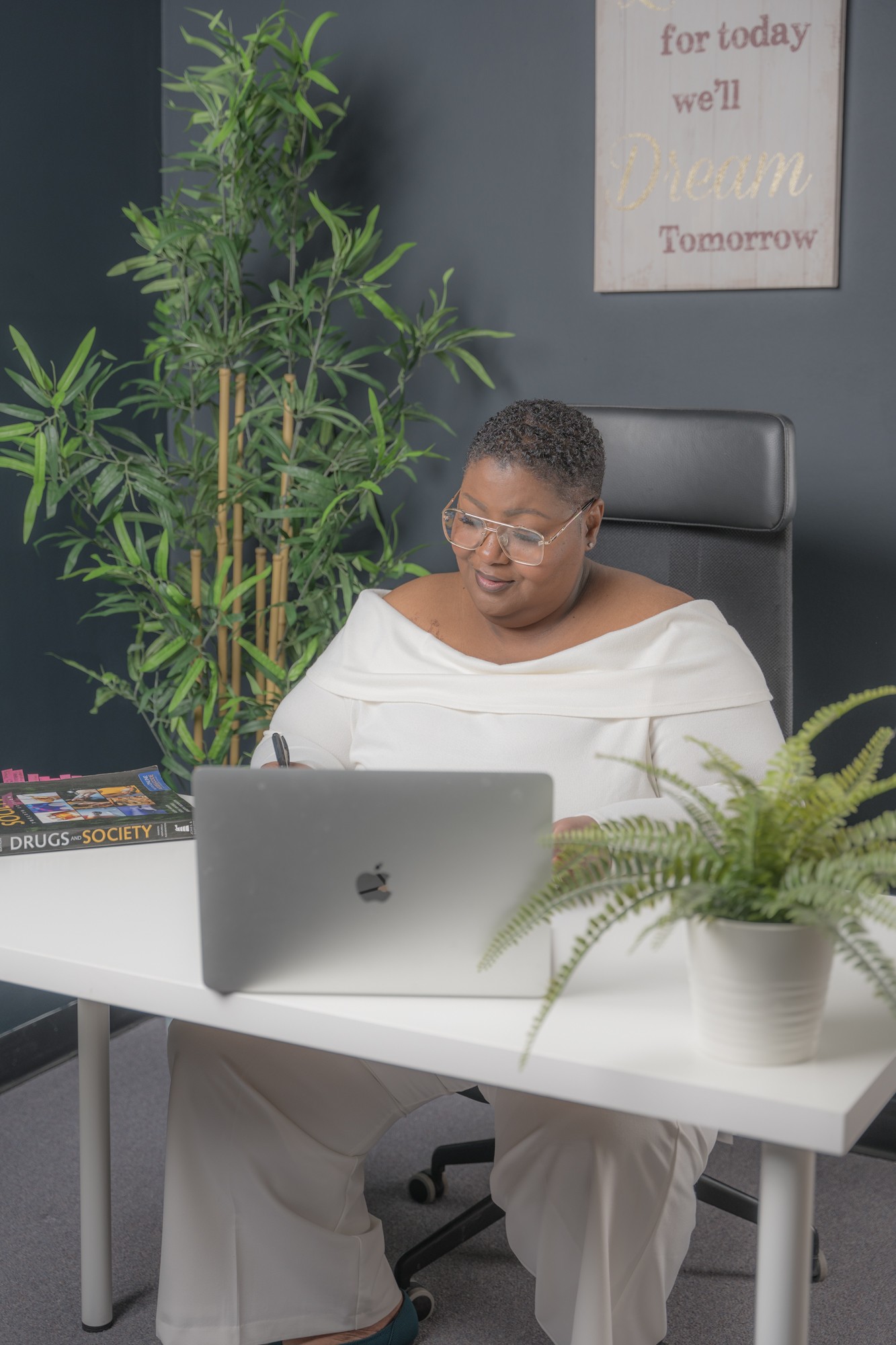 Woman sitting at a desk with a laptop, plants, and a framed quote on the wall behind her.