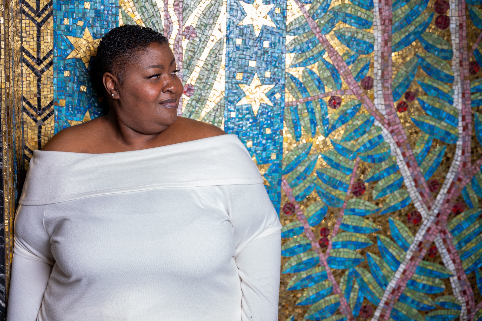 Woman with dark hair in a white off-shoulder top smiling, standing in front of a colorful mosaic background.