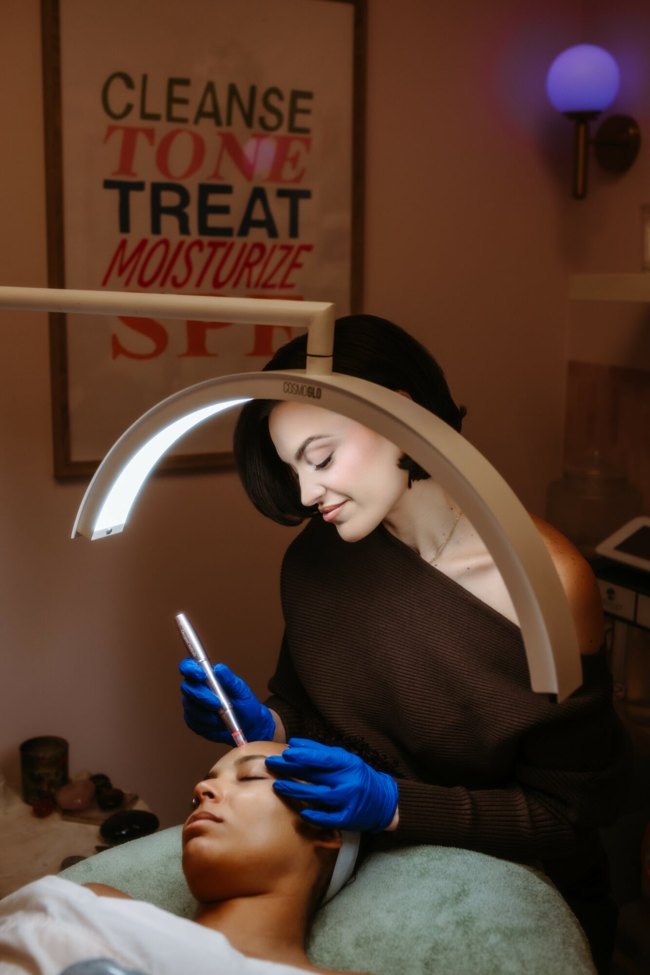A woman with black hair and blue gloves examines a person's face under a ring light in a clinical setting.