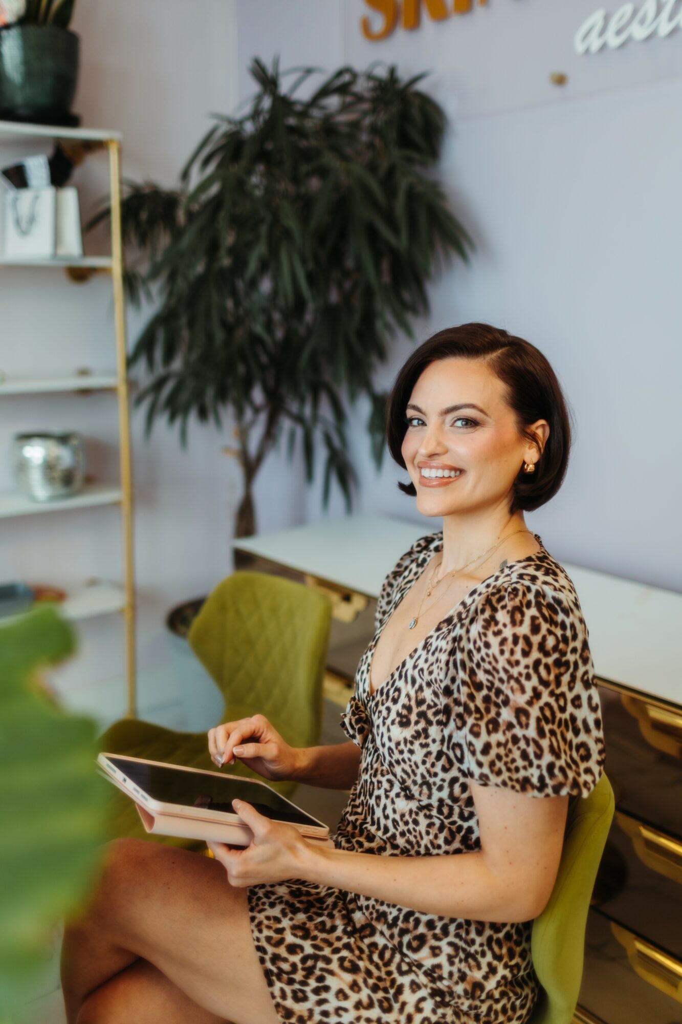 Woman with short dark hair smiling, wearing leopard print dress, holding a tablet, sitting in a modern room with plants and furniture.