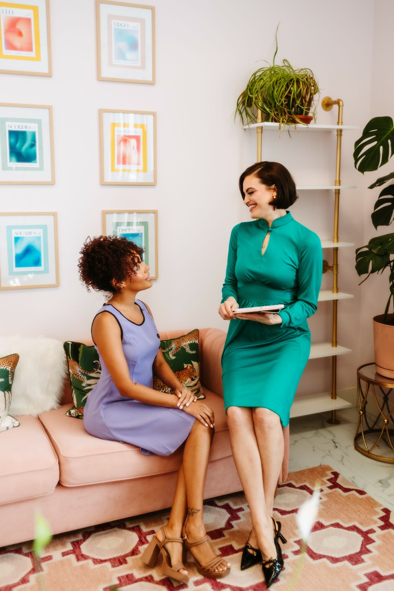 Two women talking in a living room, one sitting on a pink sofa, the other standing with a book, wall with framed art behind.