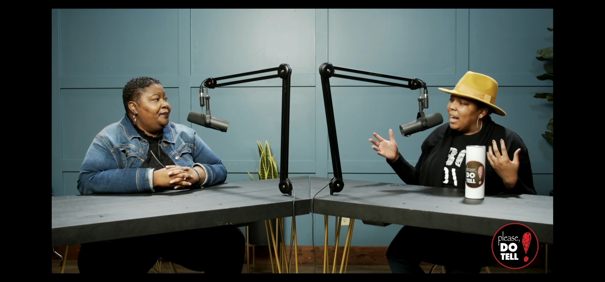 Two people sit at a table with microphones, engaged in conversation, in a recording studio setting.