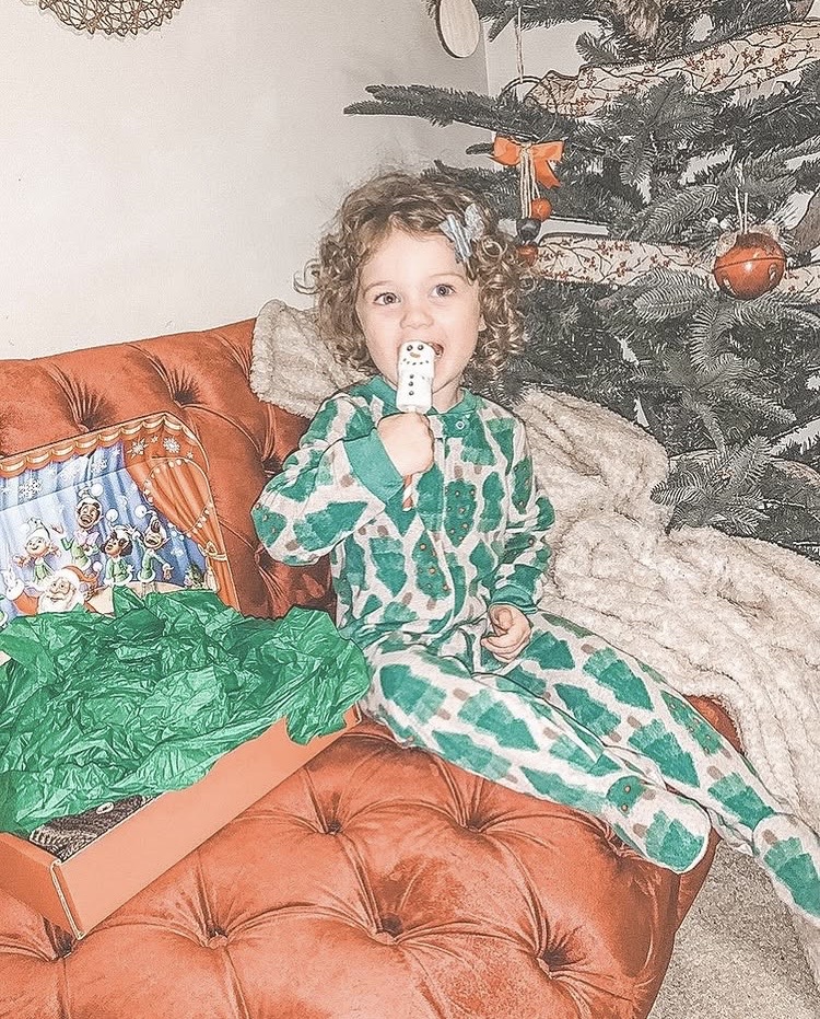 Child with curly hair in green and white pajamas sitting on a sofa, holding a remote control, Christmas tree in background.