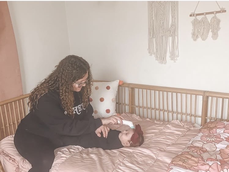 Person leaning over a child lying on a bed with a pink and brown bedspread, in a room with wall hangings.
