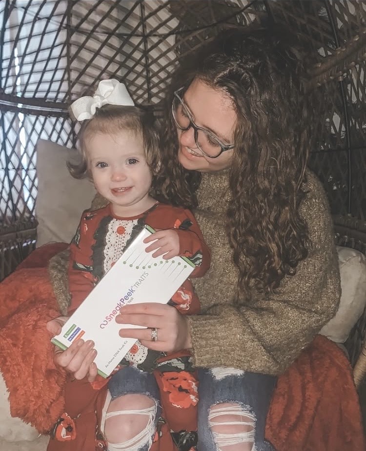 Adult woman with glasses and curly hair holding a smiling young girl with a white bow in her hair, both holding a white paper.