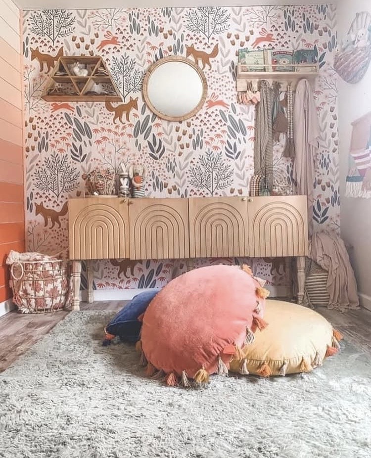 Decorative wall with animal and plant wallpaper, round mirror, shelves, and a wooden cabinet. Cushions and a rug on the floor.