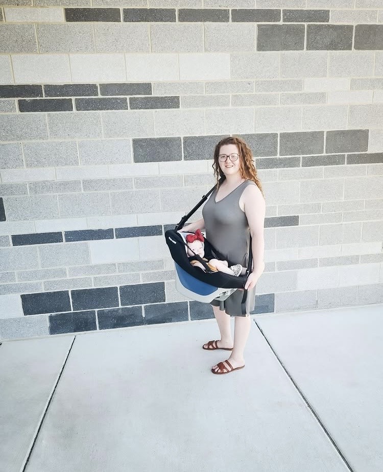 Woman standing outdoors with a bag, holding a child, against a brick wall, smiling, wearing glasses, tank top, shorts, sandals