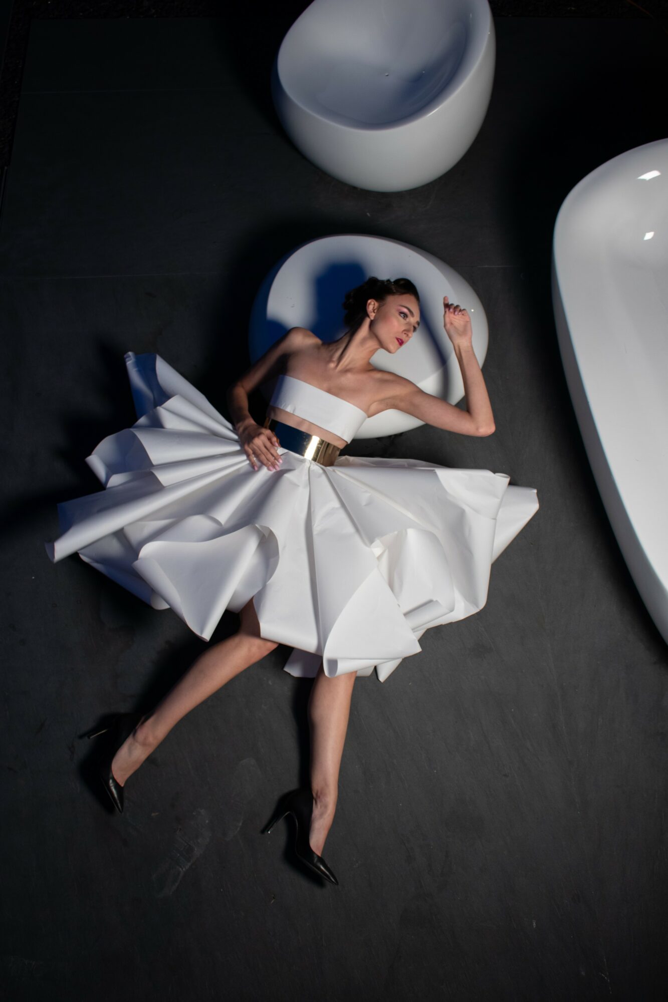 Woman in a white dress lying on the floor among large white and gray objects, with her eyes closed and arms raised.