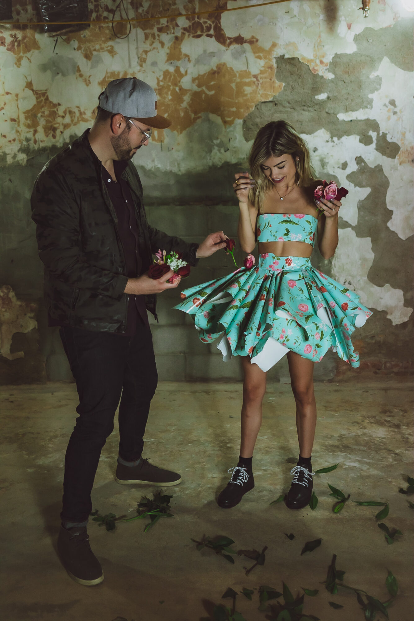 A man and a woman stand in front of a weathered wall, with butterflies on the floor and the woman holding flowers.