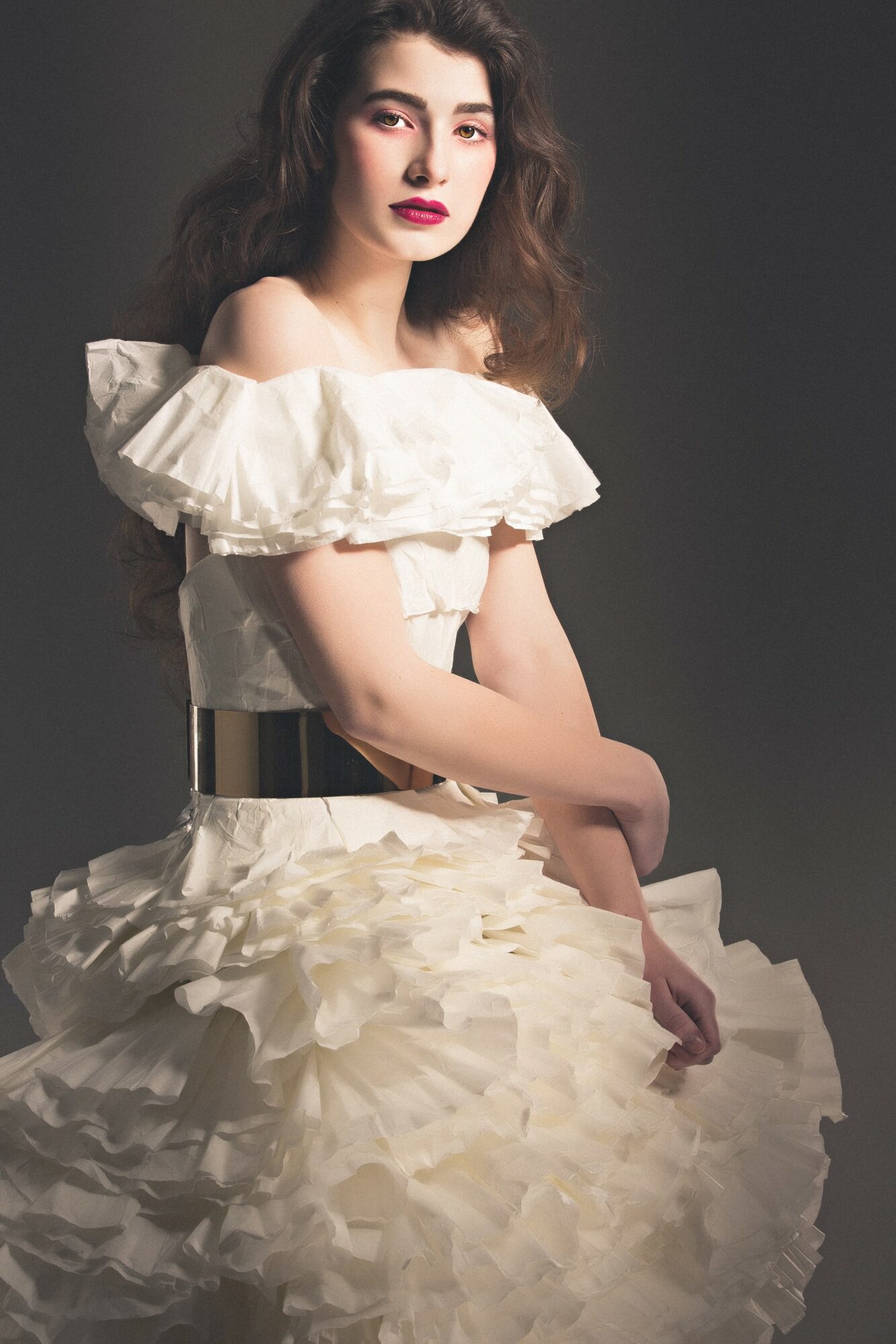 Young woman with wavy hair in a white ruffled dress with off-shoulder design, standing against a dark background.