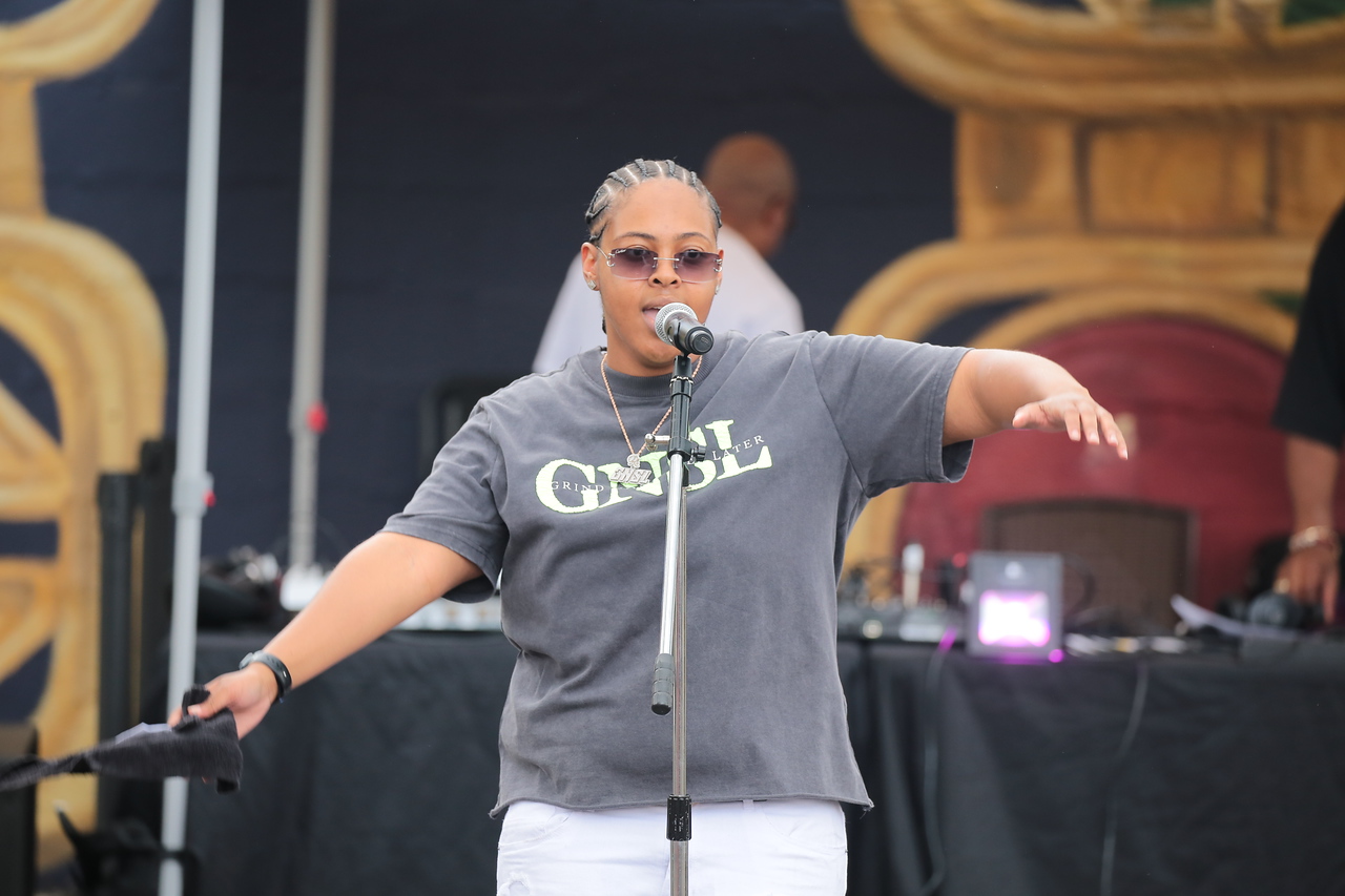Person wearing sunglasses and a gray T-shirt speaking into a microphone on stage.