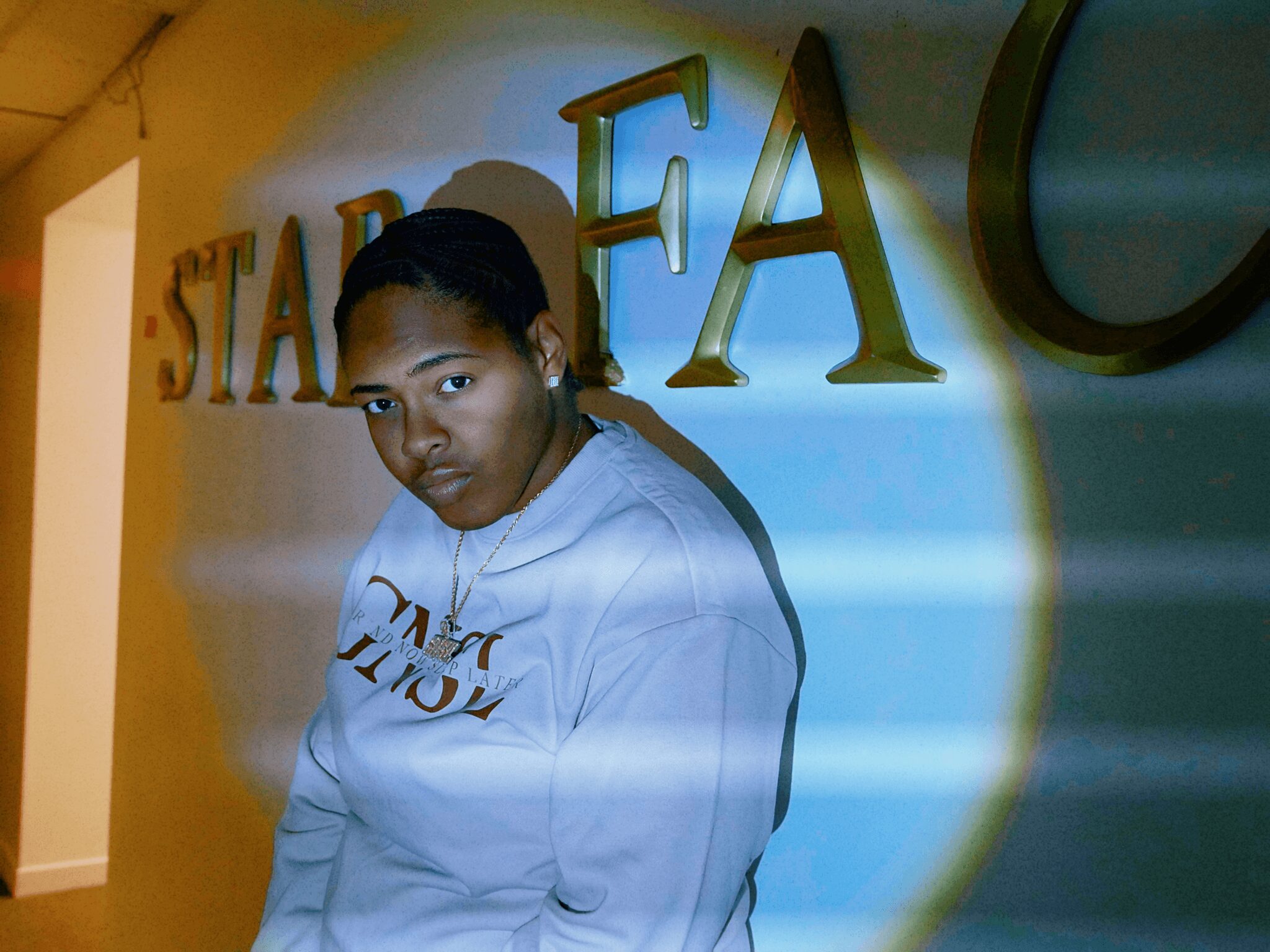 Young person with dark hair in a white hoodie standing in front of a wall with large gold letters, illuminated by a spotlight.