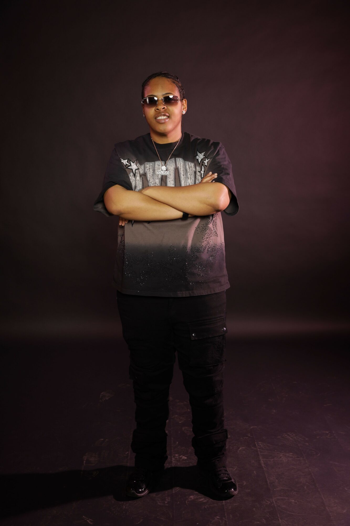 Person standing with arms crossed, wearing sunglasses and a shiny top, against a dark background.