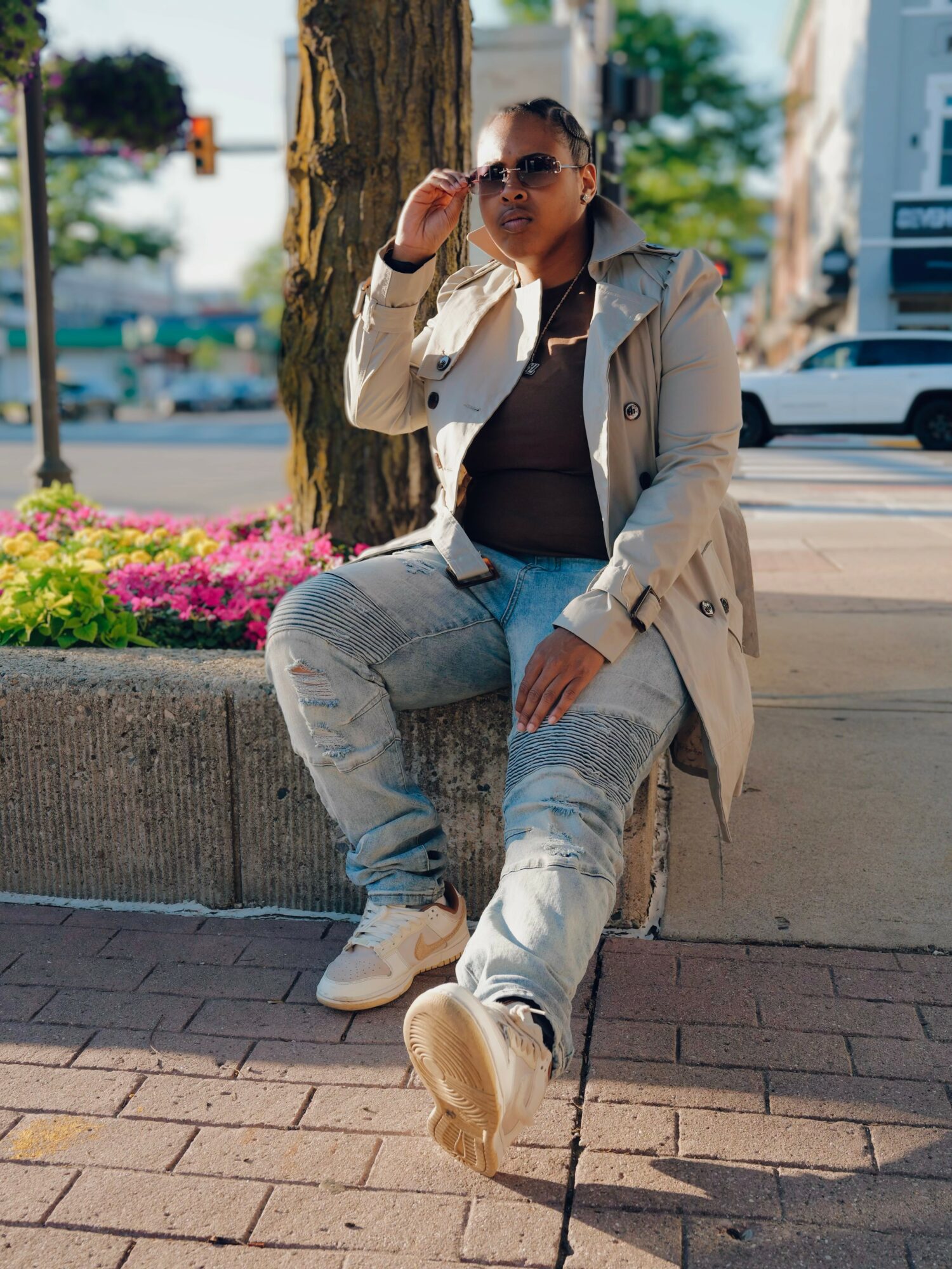 Woman sitting on a concrete ledge outdoors, wearing sunglasses, a beige coat, black top, and light jeans, with pink flowers nearby.