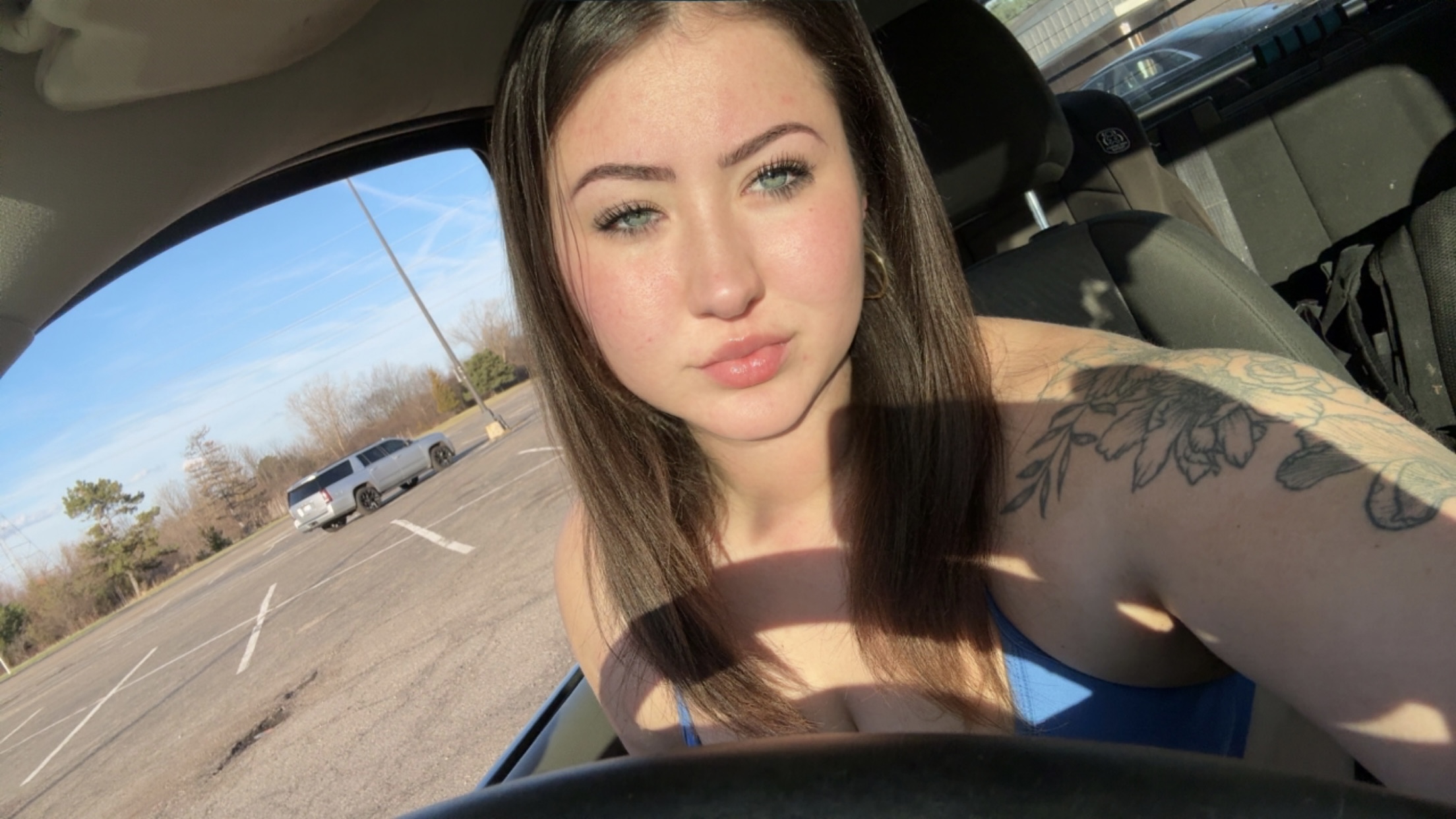 Young woman with long brown hair taking a selfie inside a car, winking and making a pouty face, with sunlight on her face.