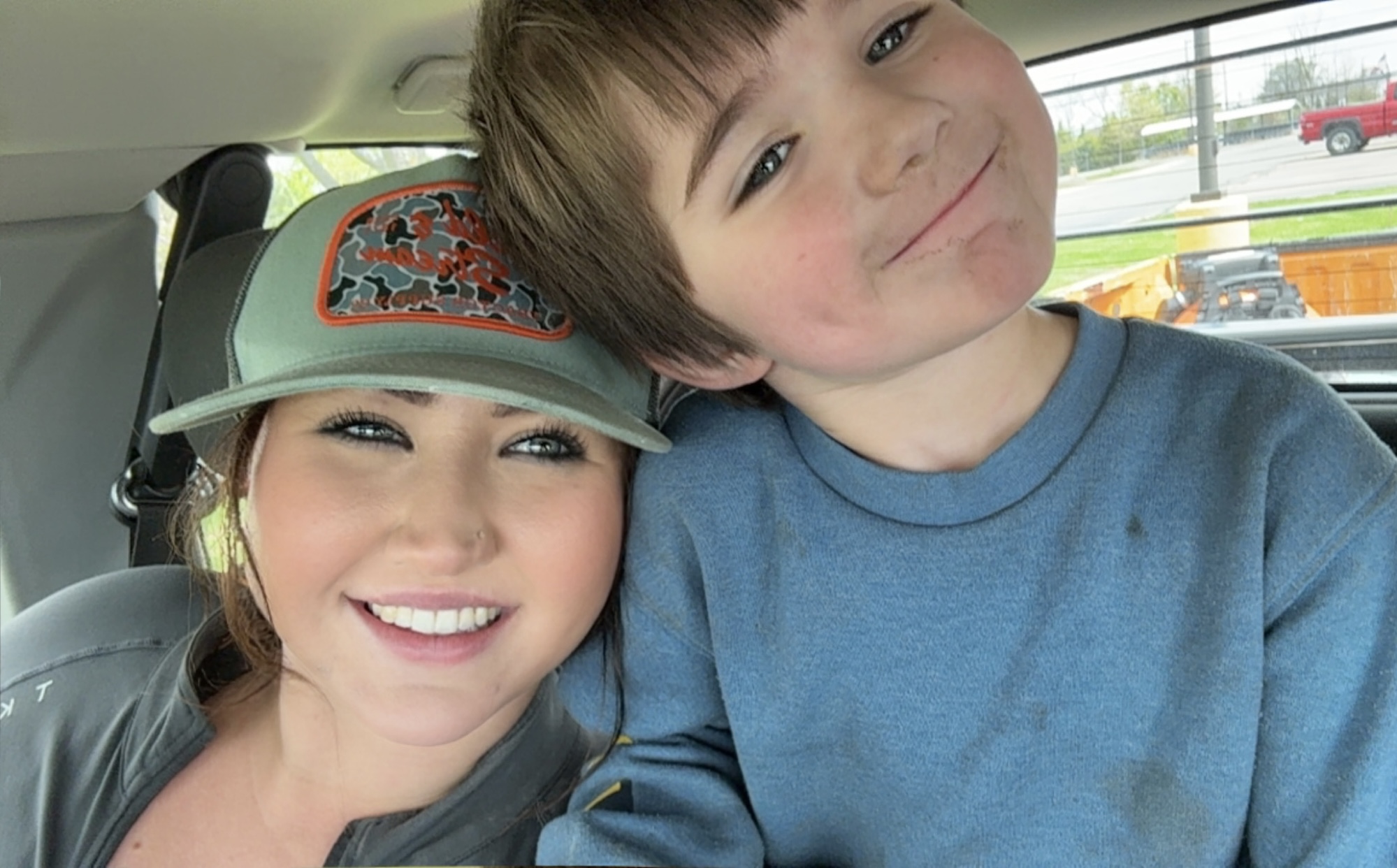A woman and a young boy smiling inside a vehicle, close together, with the woman wearing a cap.