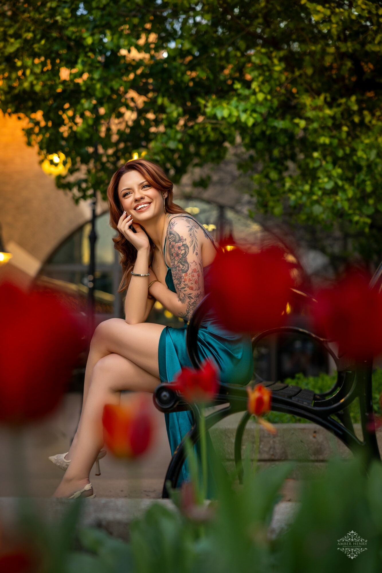 Smiling woman with tattoos sitting on a park bench, surrounded by red tulips and green trees.