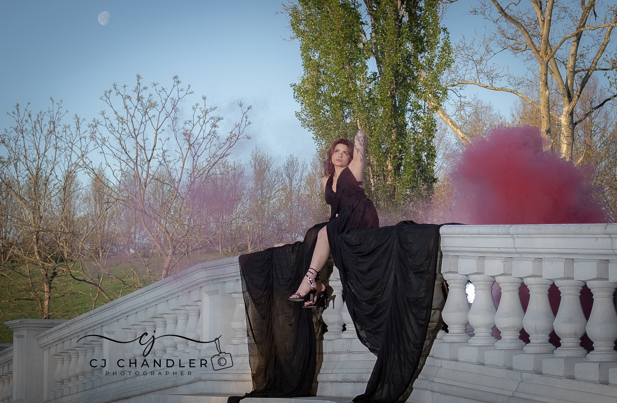 Woman in a black dress sitting on a white balustrade with pink smoke behind her outdoors, trees in the background.