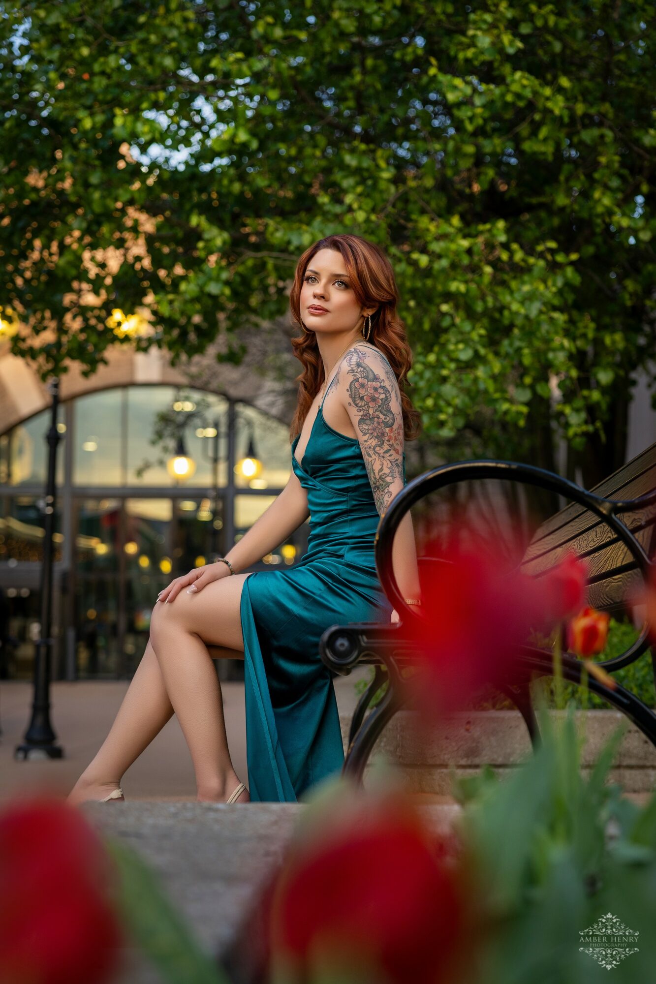 Woman with tattoos in a teal dress sitting on a bench outdoors, trees and a building in the background.