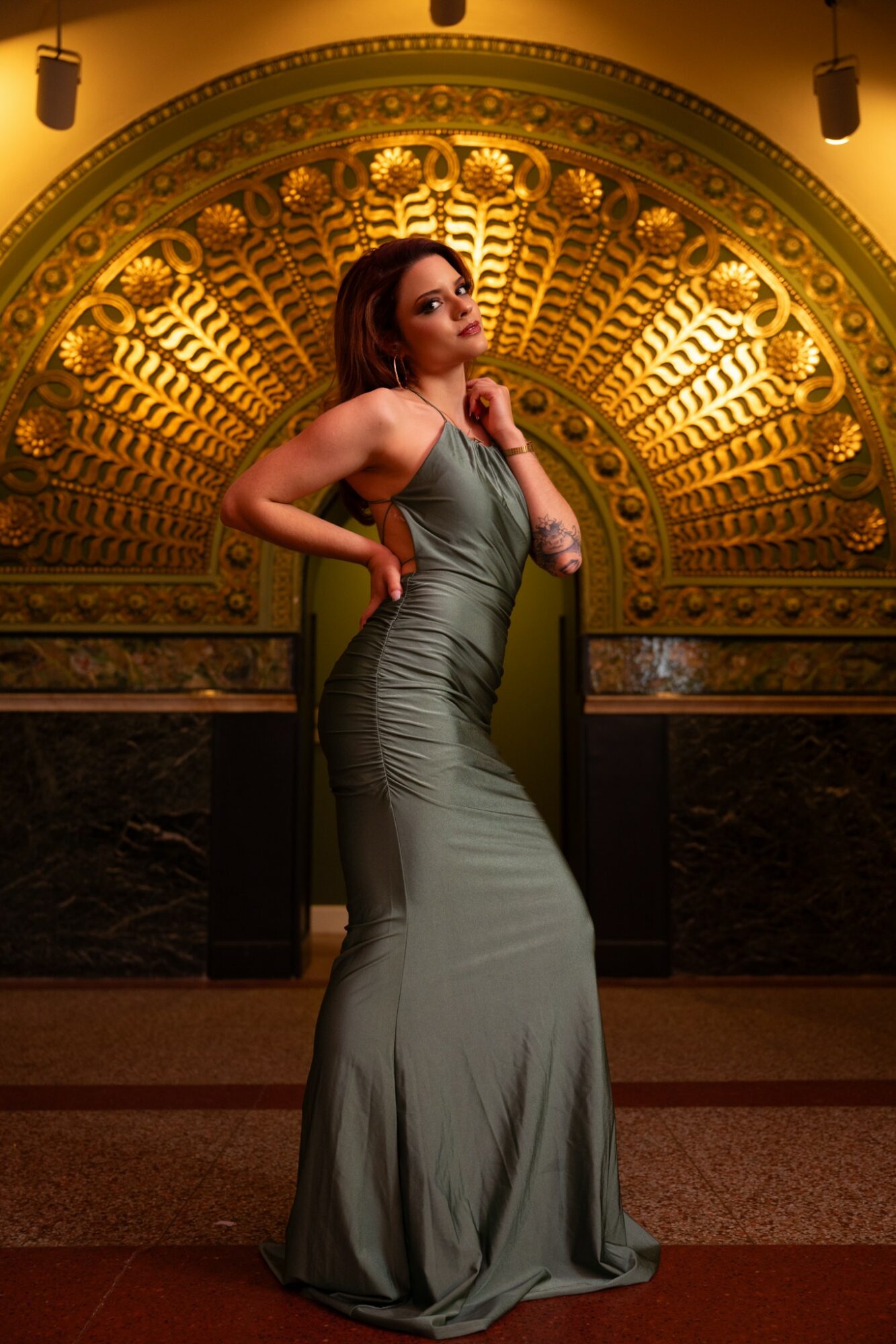 Woman in a long, elegant gown standing in front of a ornate, golden arch with intricate patterns, posing confidently.