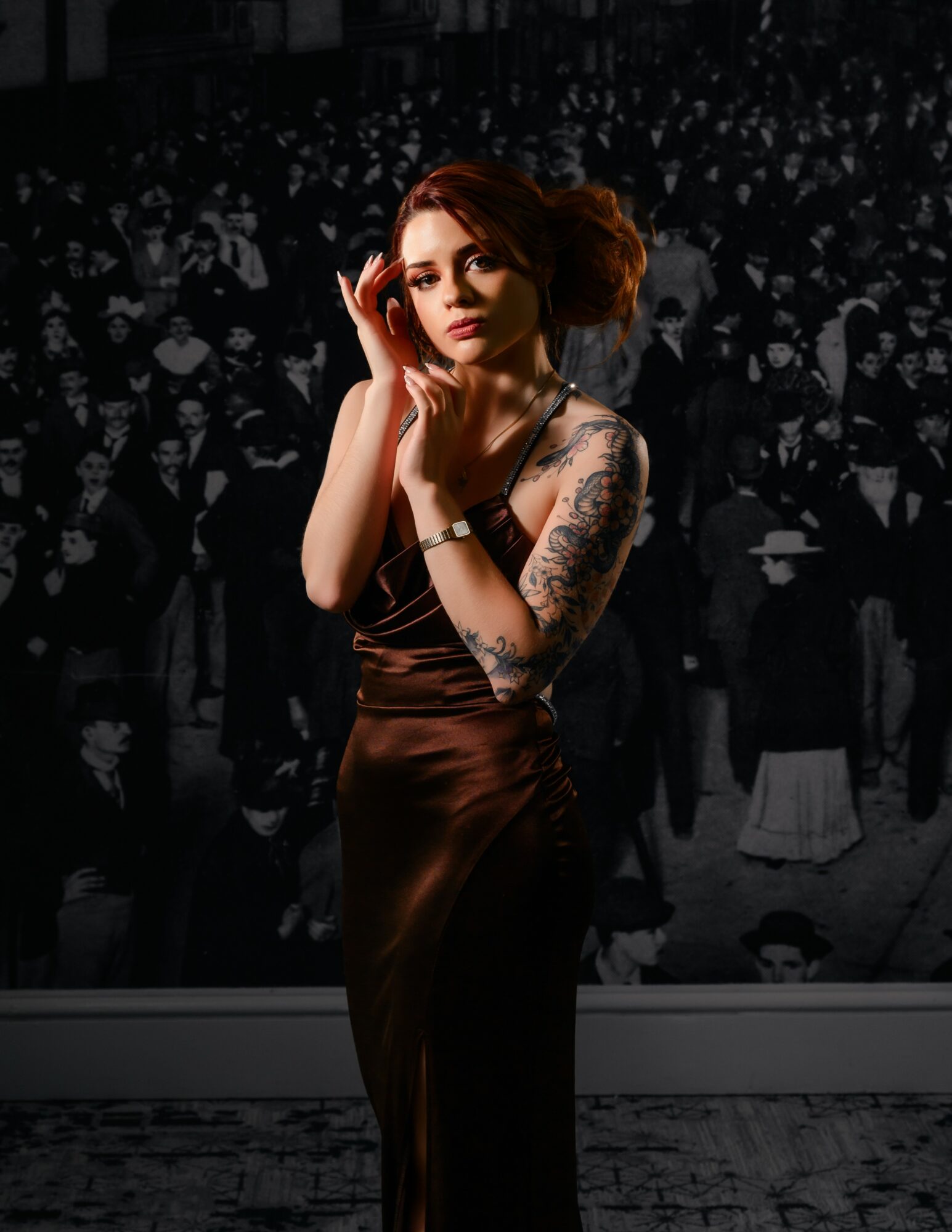 A woman with red hair and tattoos on her arms stands in front of a black-and-white crowd background, touching her face.