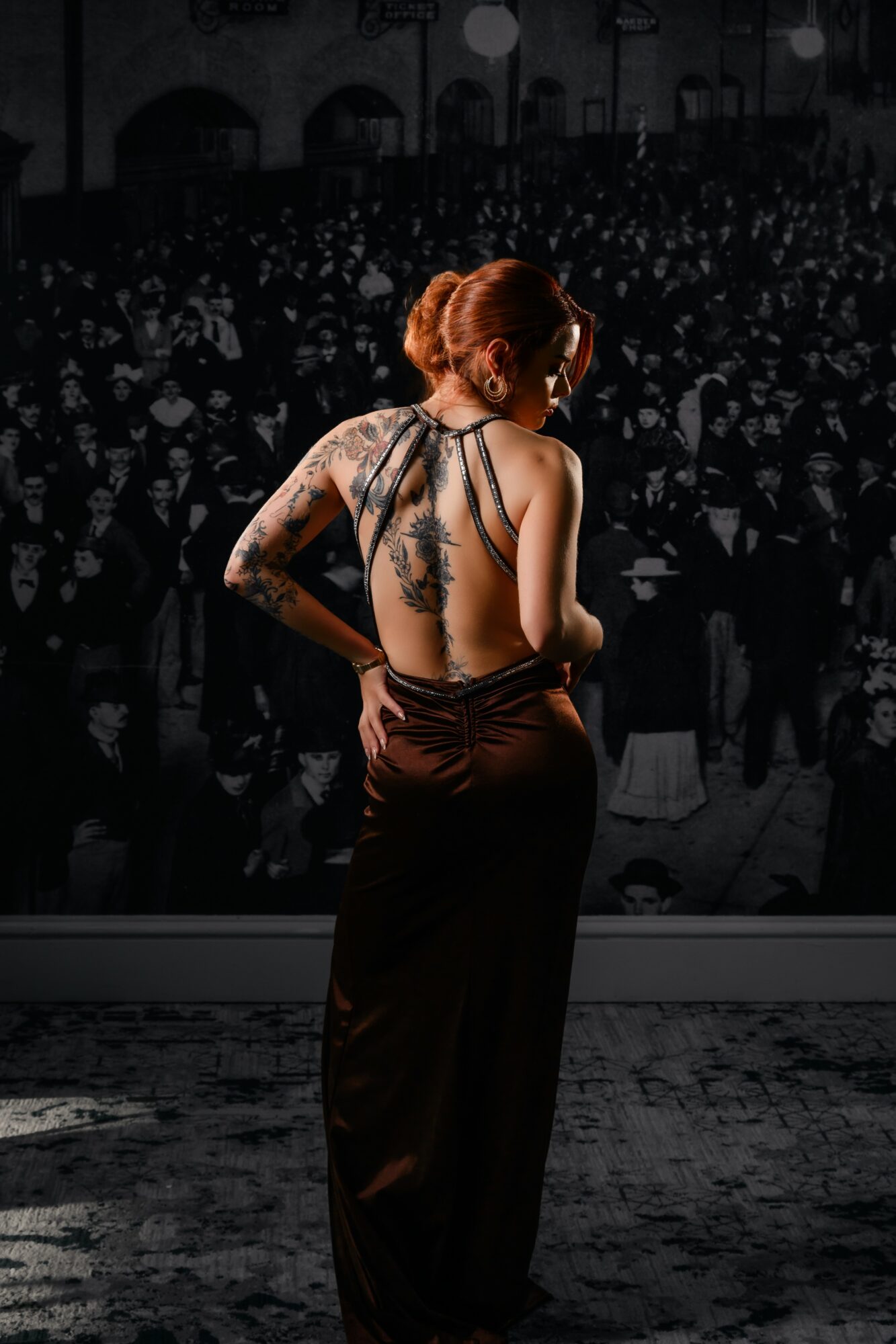 Woman with red hair and tattoos on her back, wearing a backless dress, facing a large audience in a dark venue.