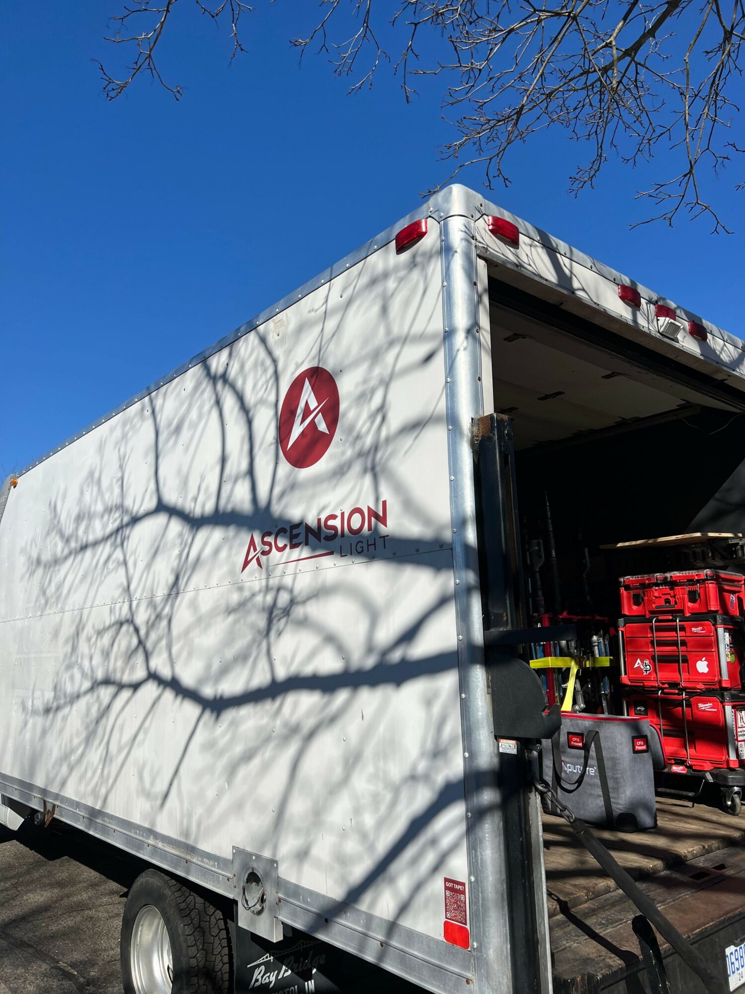 Side view of a white mobile unit with a red logo and text, shadows of tree branches on the surface.