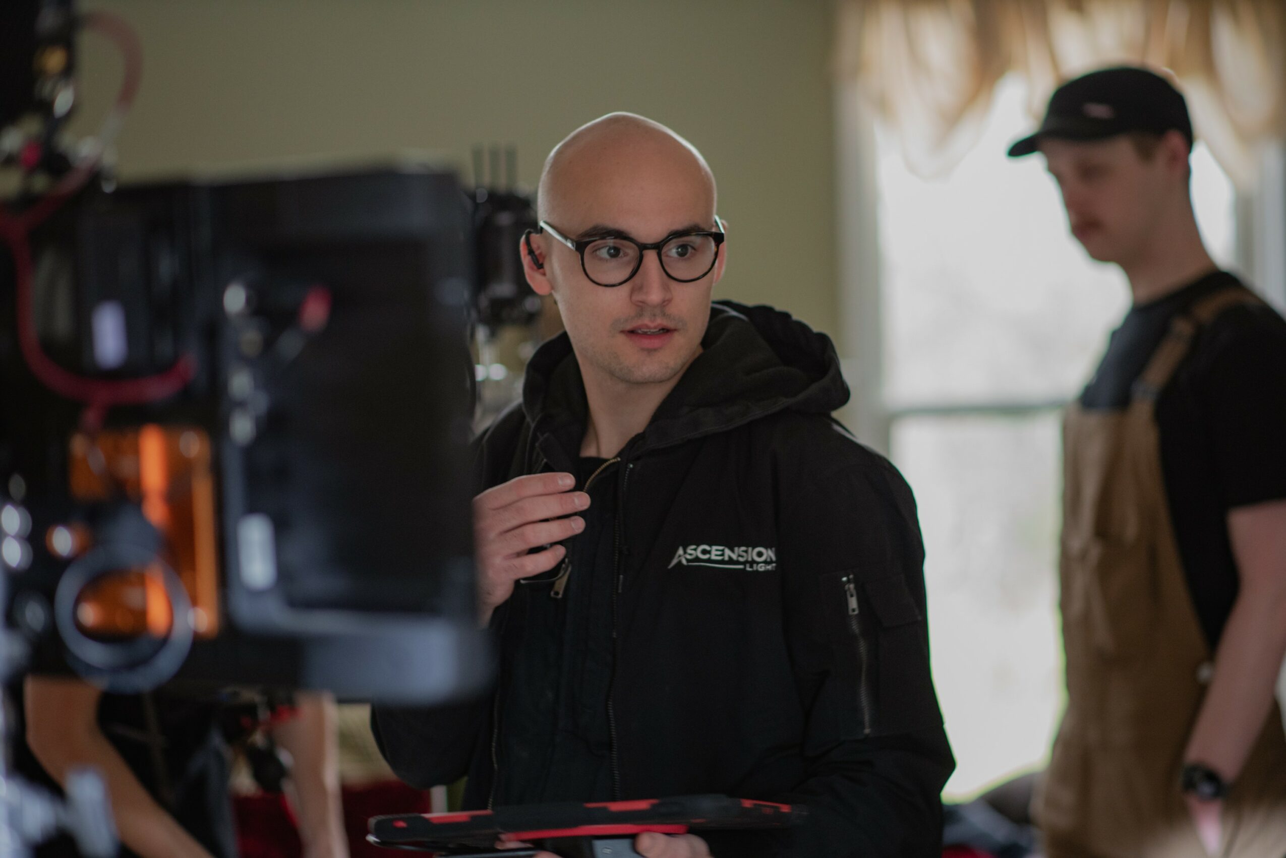 Bald man with glasses and black hoodie looking at camera, person in apron in background, indoor setting with window light
