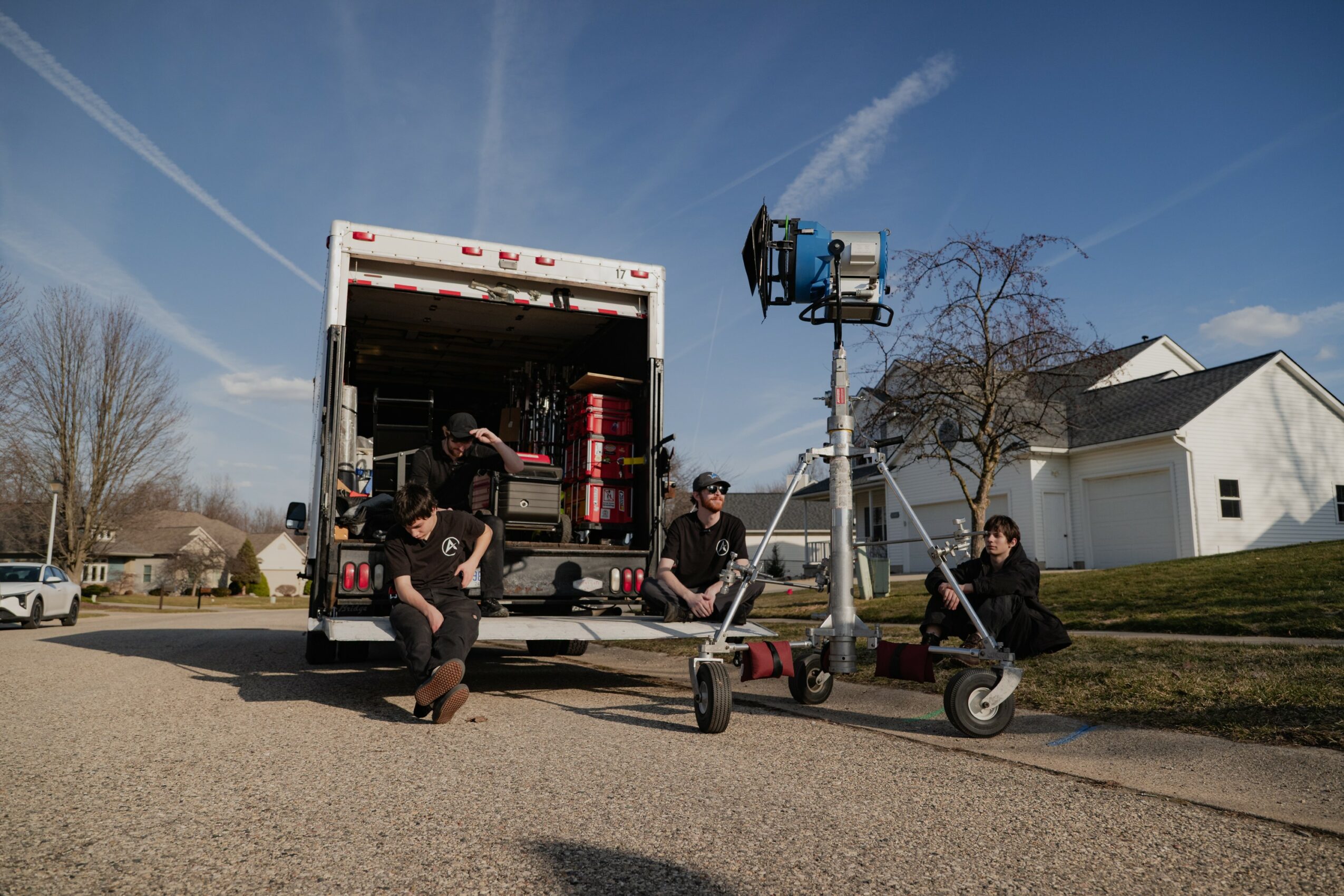 People working with equipment near a truck on a street, with a lift and a house in the background.