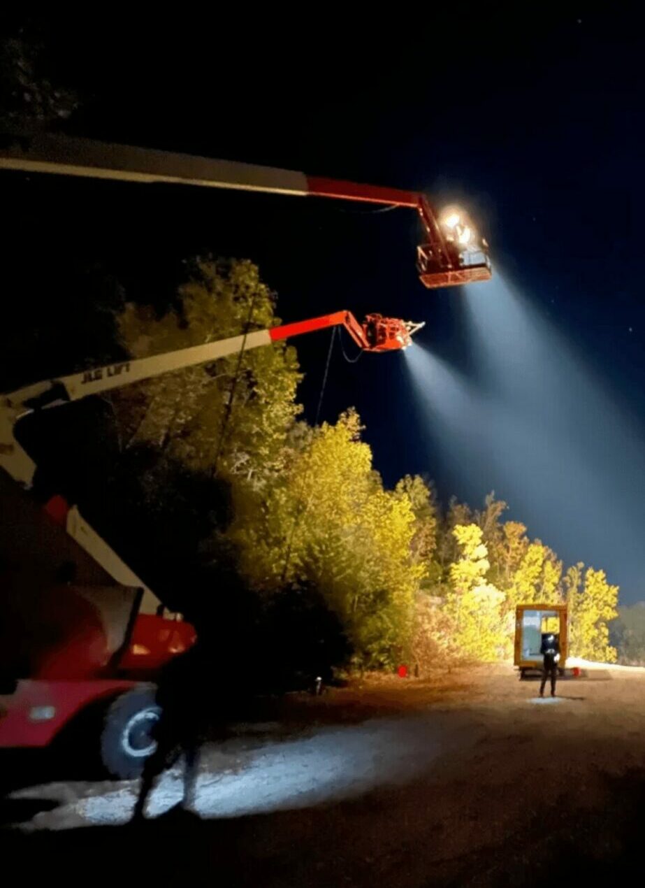 Night scene with a crane, trees, and a person holding a phone, illuminated by bright lights and a beam of light.