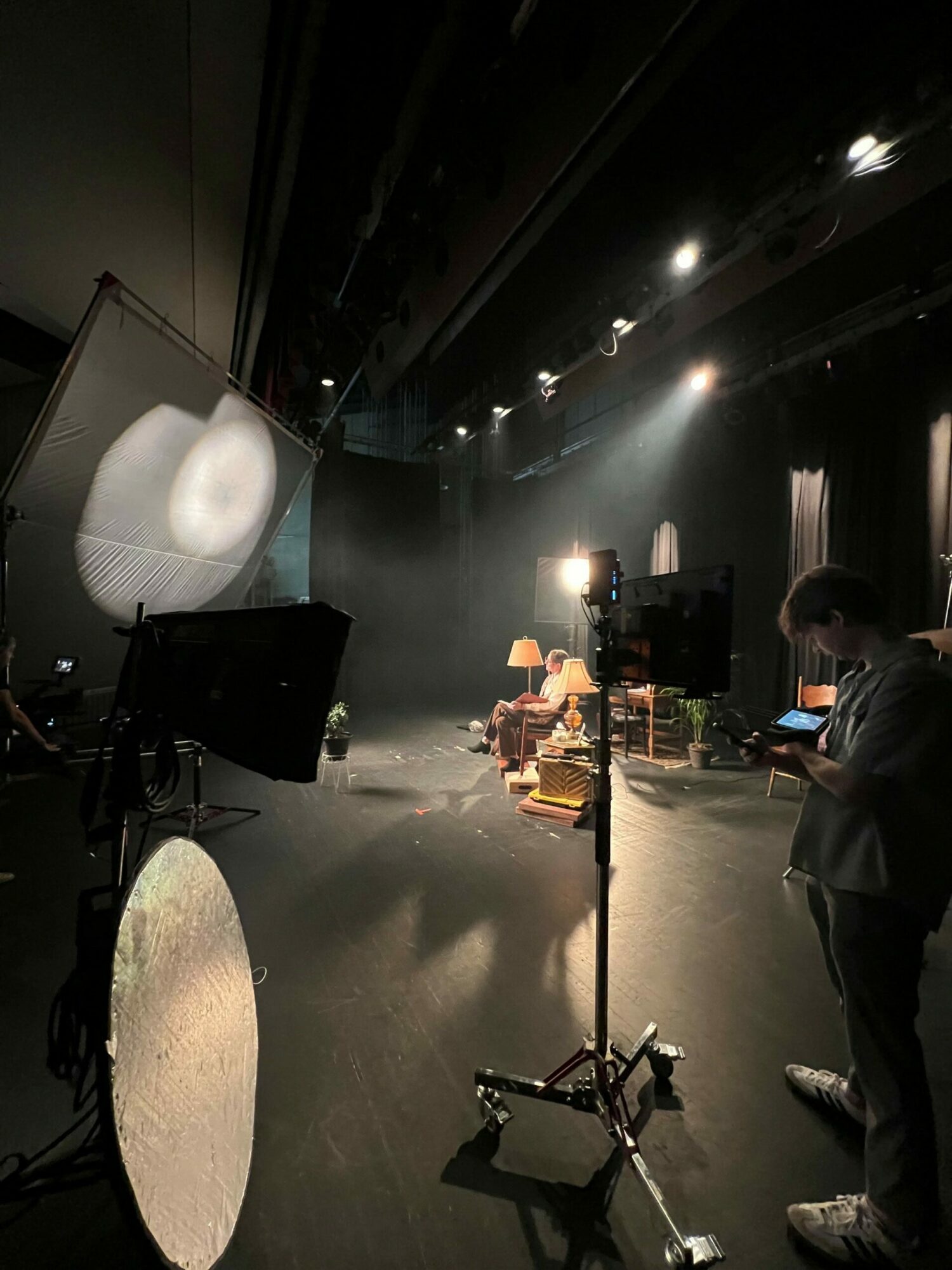 People working on a stage with equipment, lights, and a large reflector, in a dimly lit environment.
