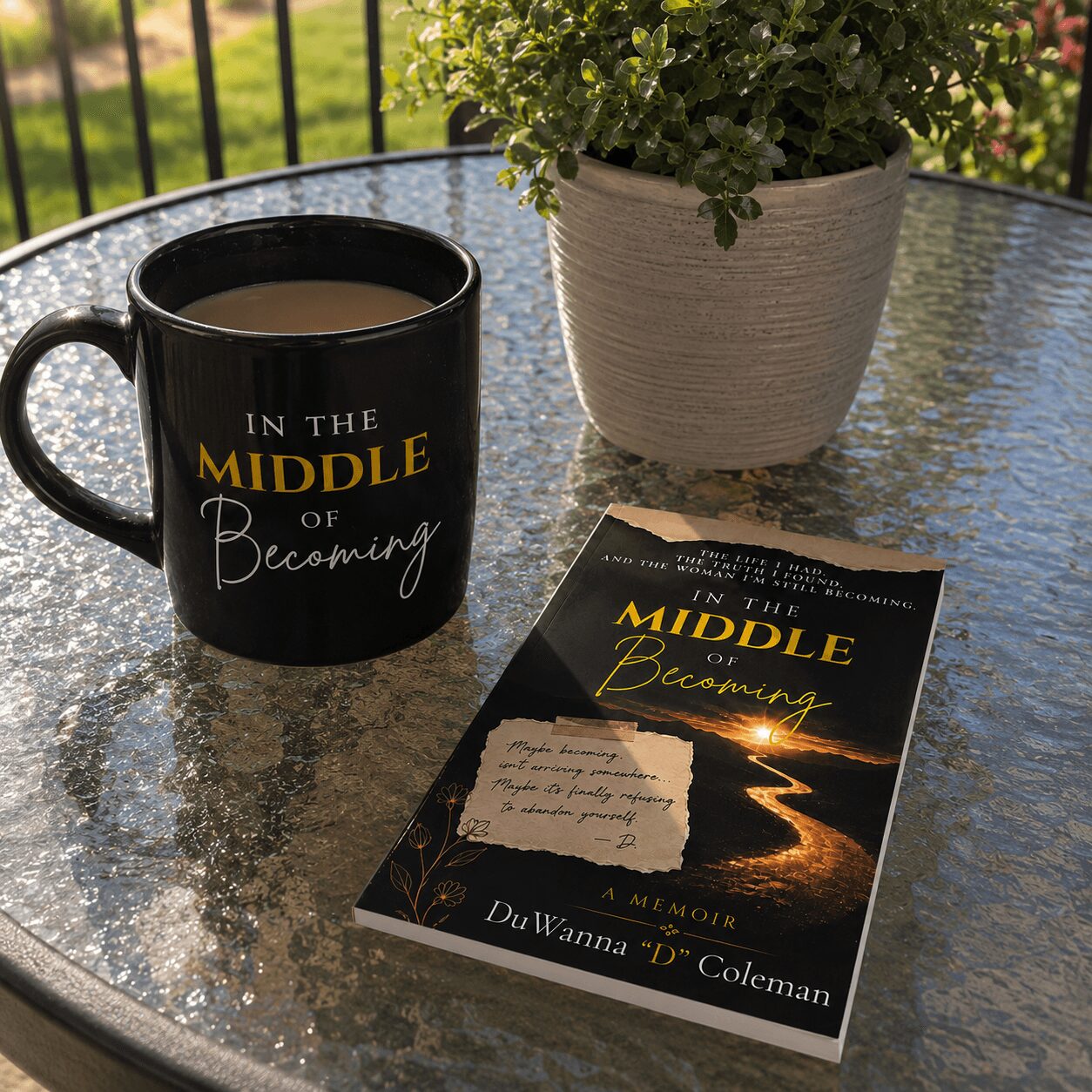 Black mug with yellow and white text, potted plant, and a book on a reflective glass table outdoors.