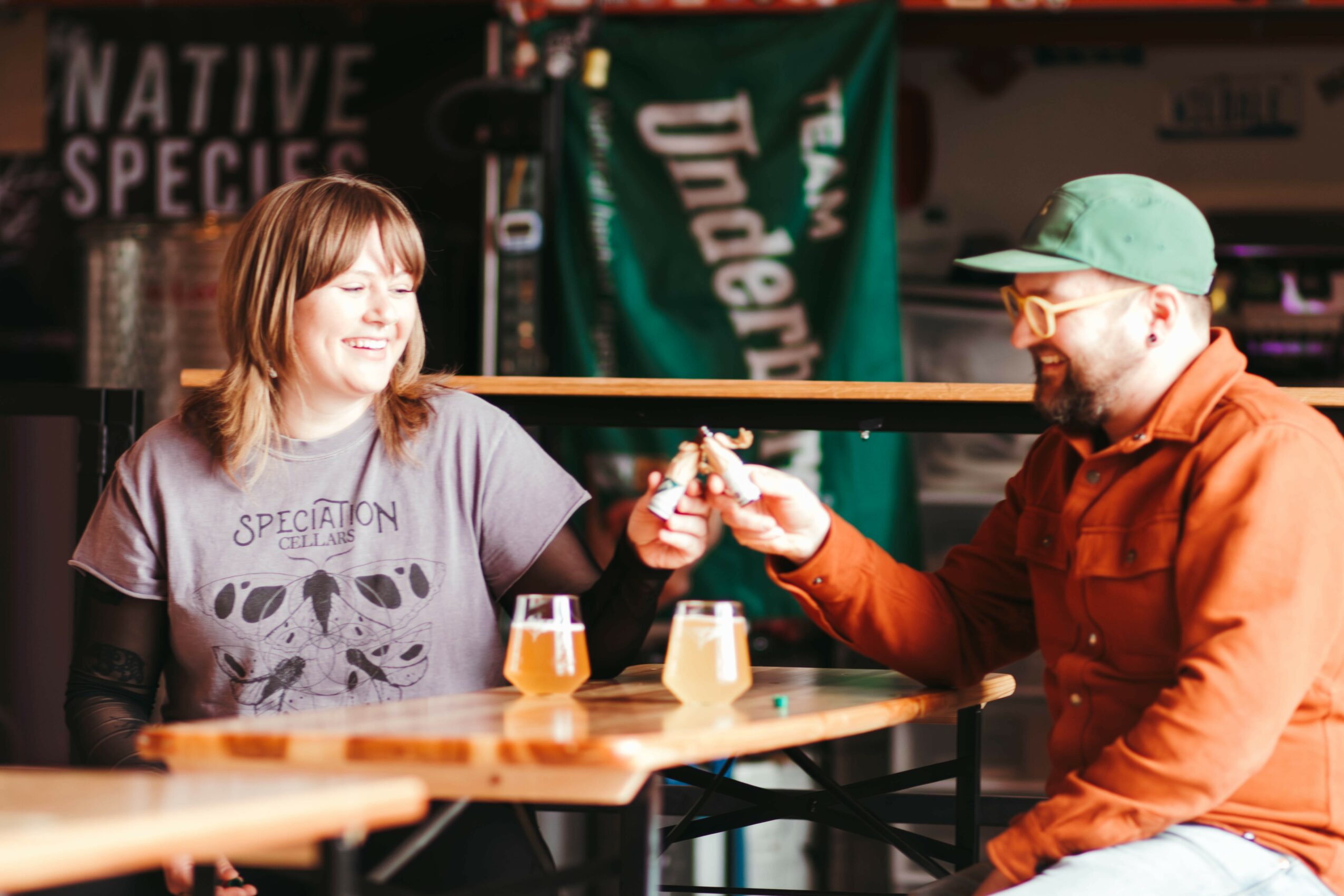 Two people smiling and exchanging a small object at a bar, with drinks on the table and banners in the background.