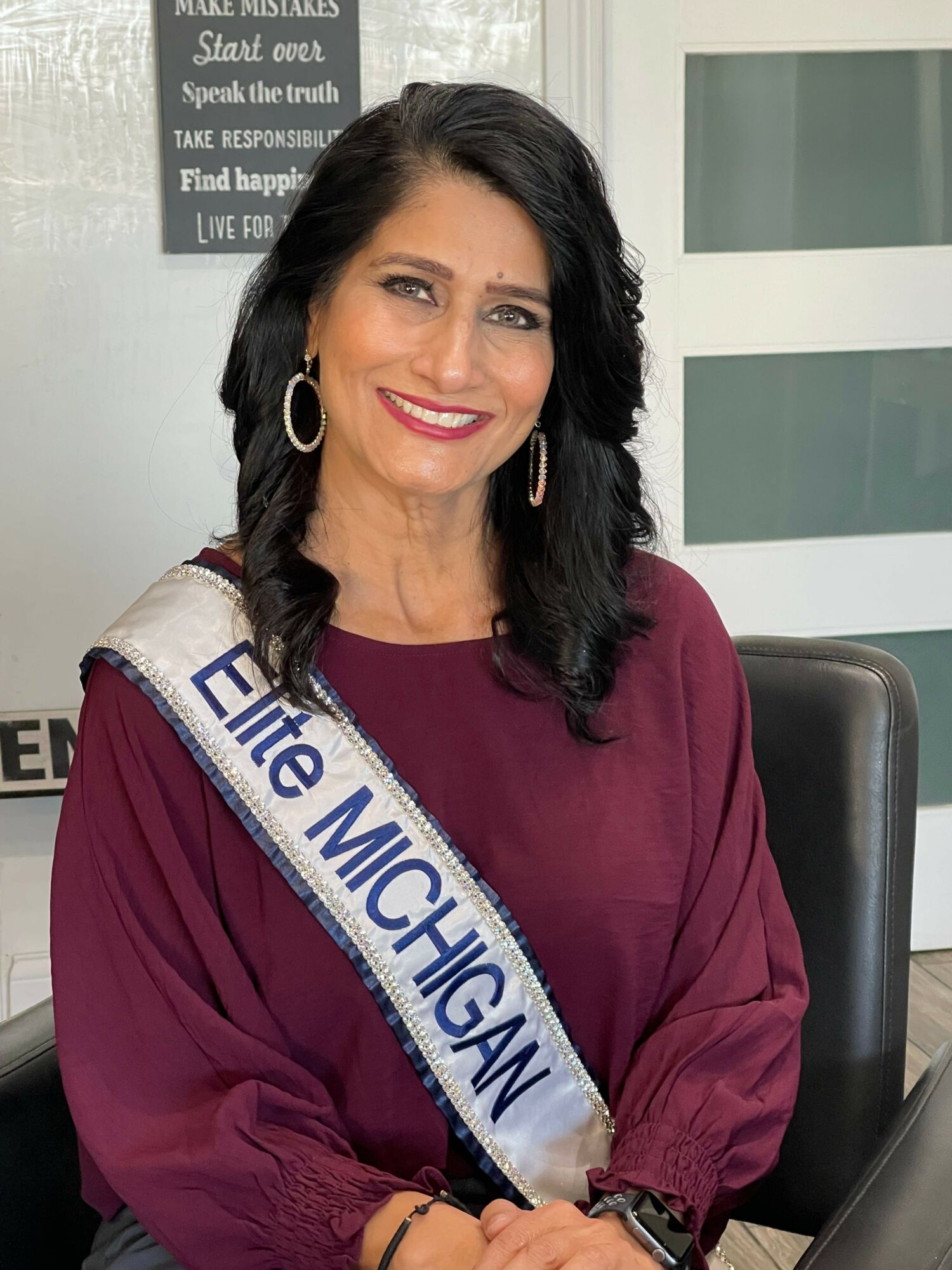 Woman with dark hair and earrings wearing a maroon top and a sash reading 'Elite MICHIGAN' sitting at a desk.