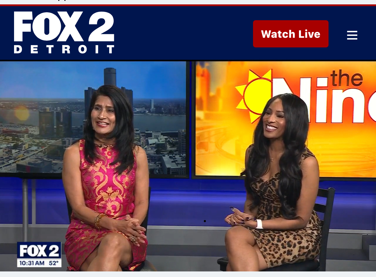 Two women sit on a news set with screens behind them displaying cityscape and logo, smiling and talking.