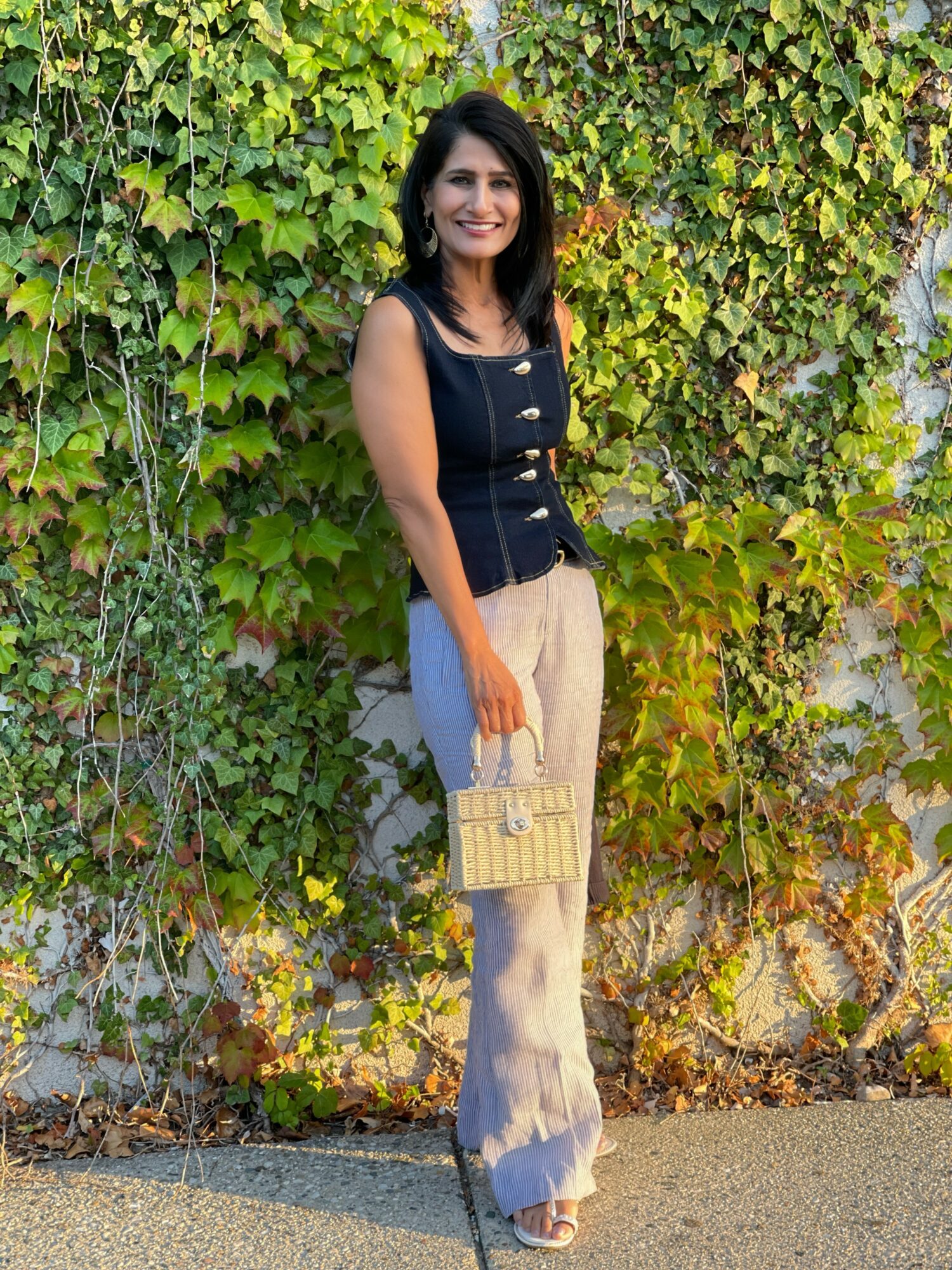 Woman standing outdoors in front of green leafy wall, smiling, holding a beige handbag, wearing a black sleeveless top and beige pants.